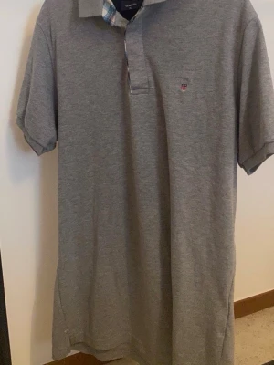 Grå pikétröja från Gant - Klassisk grå pikétröja från Gant med korta ärmar och krage. Tröjan har en liten broderad logga på bröstet och knappslå med rutigt tyg på insidan. Perfekt till en avslappnad men ändå stilren look.