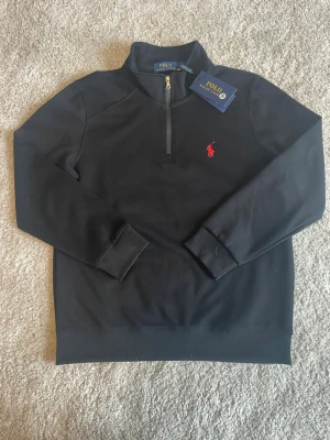 Svart långärmad polotopp Polo Ralph Lauren - Svart långärmad polotopp från Polo Ralph Lauren med klassisk röd logga på bröstet. Tröjan har en half zip-dragkedja och ribbade muddar vid ärmslut och nederkant. Perfekt för en stilren och avslappnad look.