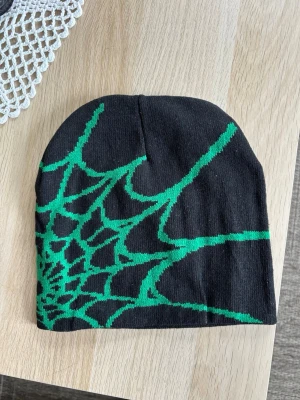 Svart mössa med grön spindelnätsdesign - Cool svart mössa med ett stort grönt spindelnätsmönster över hela framsidan. One size unisex. Jag har fler färger och skickar gärna i bundle. Mössan är mjuk och stretchig, perfekt för dig som vill sticka ut med en unik accessoar. Passar både till streetwear och casual outfits.