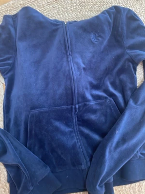 Mörkblå velourhoodie Juicy Couture - Mysig mörkblå hoodie i mjuk velour från Juicy Couture. Dragkedja framtill, känguruficka och broderad logga på bröstet. Perfekt för en chill och avslappnad stil.