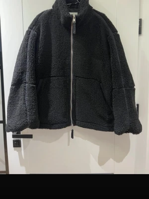 Svart oversized teddyjacka - Mysig svart teddyjacka med hög krage och dragkedja framtill. Jackan har två stora fickor och en oversized passform som ger en avslappnad look. Tillverkad i fluffigt teddy-material som håller dig varm och trendig.