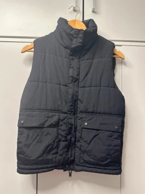 Svart dunväst med stora fickor - Snygg svart dunväst med hög krage och två stora fickor framtill med tryckknappar. Västen har quiltad design och dragkedja med vindslå. Perfekt för lager-på-lager och streetwear-stil.