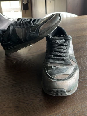 Valentino Garavani camo sneakers - Snygga sneakers från Valentino Garavani med svart och grått camomönster. Skorna har svarta snören, låg profil och detaljer i mocka och textil. Perfekta för dig som vill ha en unik och trendig look.