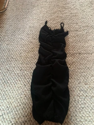 Svart kort klänning med snörning - Snygg svart kort klänning med smala axelband och trendig snörning framtill. Klänningen har en åtsittande passform och rynkade detaljer längs hela plagget. Perfekt för dig som gillar en stilren och modern look.