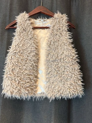 Beige fluffig pälsväst - Supersnygg beige pälsväst med fluffig, långhårig fuskpäls och silkeslen insida. Västen är ärmlös och har en öppen front, perfekt för att lyfta vilken outfit som helst. Passar dig som vill ha en trendig och mysig look.