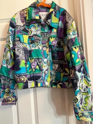 Färgstark jeansjacka från Laurèl escada - Unik vintage  jacka från Laurèl i storlek 38 med färgstarkt patchwork-mönster i lila, turkos, limegrönt och svart. Jackan har klassisk krage, två bröstfickor med lock och knappar framtill. Perfekt för dig som vill sticka ut med en kreativ och lekfull stil.