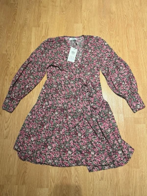 Blommig omlottklänning från Only - Kort omlottklänning från Only med blommigt mönster i rosa, vitt och grönt. Klänningen har långa ärmar, volangdetaljer och knyt i midjan. Nypris: 450 kr