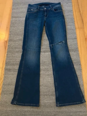 Blå bootcut jeans från Pepe Jeans - Säljer ett par blå bootcut jeans från Pepe Jeans med slitning och hål på ena låret. Klassisk femficksmodell med knapp och dragkedja. Jeansen har en snygg tvättad look och är tillverkade i denim.