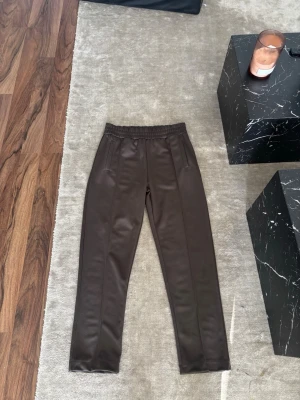 Bruna mjukisbyxor från Palm Angels - Säljer ett par bruna mjukisbyxor från Palm Angels med resår i midjan och raka ben. Byxorna har fickor fram och en stor vit Palm Angels-logga på baksidan. Materialet är glansigt och ger en sportig vibe.