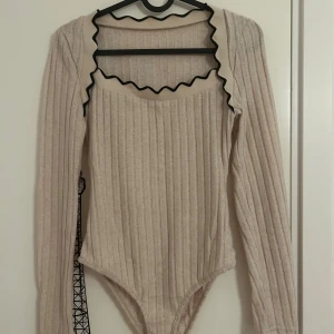 Beige ribbad långärmad body från SHEIN - Beige ribbad body med lång ärm och snygg vågig kant i svart runt halsringningen från SHEIN. Bodyn har en figurnära passform och är tillverkad i ett mjukt, stretchigt material som känns skönt mot huden. 