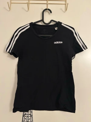 Svart Adidas t-shirt med vita ränder - Svart t-shirt med klassiska vita ränder på axlarna och logga på bröstet från Adidas i storlek XS. T-shirten har rund halsringning och korta ärmar. 