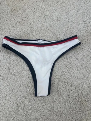 Vit bikiniunderdel med marinblå kant - Snygg vit bikiniunderdel med marinblå kant och en röd detalj längs midjan. Klassisk trosa-modell med sportig känsla och stretchigt material som sitter skönt. Perfekt för strandhäng eller poolen.