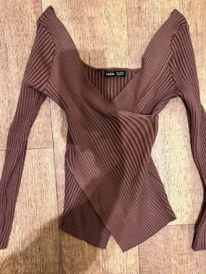 Vår Brun omlottopp  - Snygg brun omlottopp  med ribbad struktur och v-ringad hals. Toppen har långa ärmar och en asymmetrisk nederkant som ger en unik look. Perfekt för dig som vill ha något trendigt och bekvämt.