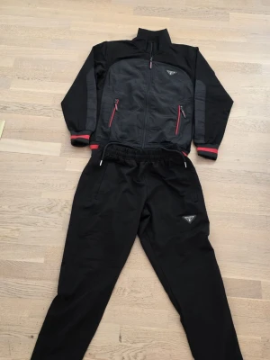 Svart träningsset från Head - Snyggt svart träningsset från prada med jacka och byxor. Jackan har dragkedja, två fickor med röda detaljer och grå paneler på ärmarna. Byxorna har resår i midjan och prada-logga på benet. Perfekt för chill eller träning.