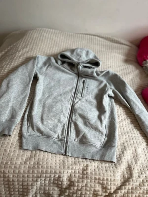 Grå hoodie från Sail Racing - Snygg ljusgrå hoodie från Sail Racing med dragkedja framtill och en extra bröstficka med zip. Tillverkad i mjukt bomullsmaterial och har en klassisk huva. Perfekt för chill dagar eller när du vill ha en clean look.