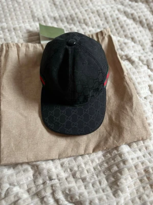 Svart keps från Gucci med GG-mönster - Snygg svart keps från Gucci med klassiskt GG-monogram över hela kepsen. På sidan finns en grön och röd rand som ger den en ikonisk look. Tillverkad i canvasmaterial och levereras med originalpåse och taggar.