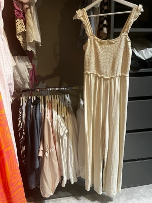 Krämvit jumpsuit med smock - Supersöt krämvit jumpsuit med smockad överdel och breda axelband med volangdetaljer. Vida ben och mjukt, luftigt tyg som känns perfekt för sommaren. Ingen synlig logga eller märke. Passar dig som gillar en avslappnad men ändå söt stil.