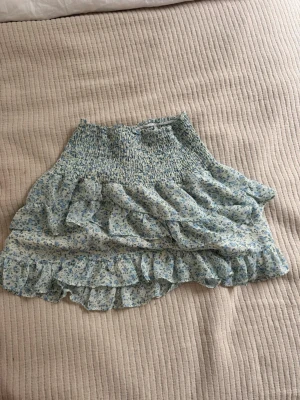 Blommig volangkjol från Neo Noir - Supersöt kort kjol från Neo Noir med blått och grönt blommönster på vit botten. Kjolen har smock i midjan och flera volanger. Helt nytt skick.