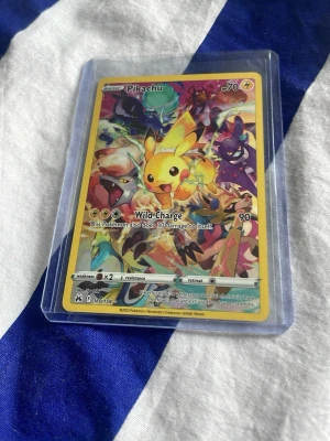 Pokémonkort Pikachu Vivid Voltage - Säljer ett färgglatt Pokémonkort med Pikachu från Vivid Voltage-serien. Kortet har en gul ram och motiv med flera Pokémon i bakgrunden. Det är tillverkat i laminerad papp och ligger i ett genomskinligt plastfodral.