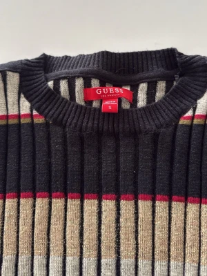 GUESS Randig ribbstickad tröja - Snygg ribbstickad tröja från GUESS i storlek S. Tröjan har långa ärmar och ett randigt mönster i fina färger som är lätt att kombinera med allt  (svart, beige, grått, vinrött och guld). Rund halsringning och figurnära passform. Snygga guldiga knappar ger en exklusiv känsla. Perfekt för dig som gillar trendiga och färgglada plagg.
