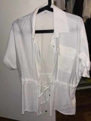 Vit skjorta med snörning och ficka - Vit skjorta i mjukt material med korta ärmar, snörning i midjan och en bröstficka. Skjortan har klassisk krage och knappar framtill. 