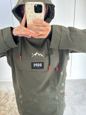 Olivgrön Dope Snow vindjacka - Snygg olivgrön vindjacka från Dope Snow med stor magficka, huva och svarta dragskor med orange detaljer. Jackan har ett vitt bergstryck på bröstet och patch på ärmen. Materialet är vattenavvisande och perfekt om du åker mycket skidor, den är i strl XS men skulle säga att den känns mer som en S.