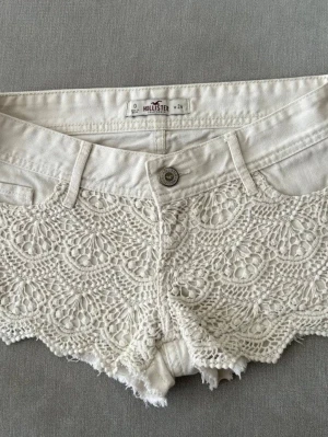 Vita spetsdetaljerade shorts Hollister - Säljer ett par vita shorts från Hollister med snygga spetsdetaljer framtill och klassisk femficksdesign. Shortsen har låg midja och dragkedja framtill. Perfekta för sommarens alla äventyr!