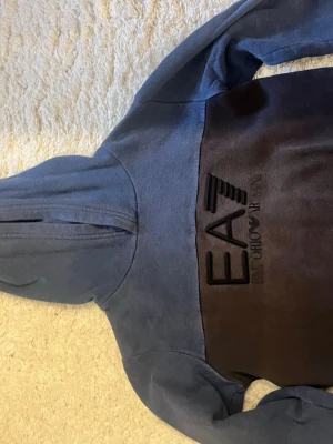 Blå EA7 Emporio Armani hoodie - Snygg blå hoodie från EA7 Emporio Armani med stor logga på bröstet. Hoodien har huva och är gjord i mjukt material som känns skönt mot huden. Perfekt för dig som gillar streetwear och vill ha något stilrent men ändå sportigt.