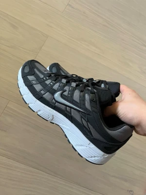 Nike svart/grå mesh sneaker - Snygga sneakers från Nike i svart och grått med silvriga detaljer. Ovandelen är i mesh och syntet för bra andningsförmåga och komfort. Klassisk Nike Swoosh på sidan och vit, rejäl sula för skön dämpning. Perfekta för dig som gillar sportig stil.