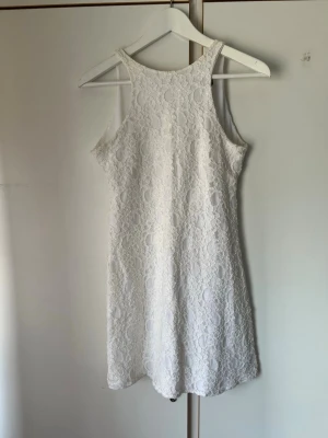 Vit spetsklänning med knappar - Söt vit kortklänning i spets med rund halsringning och ärmlös design. Klänningen har fina spetsdetaljer över hela tyget och knappar i ryggen som ger en elegant touch. Perfekt för sommarens alla tillfällen.