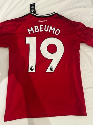 Manchester United, Mbeumo 19, 25/26 - Manchester United hemmtröja 25-26 från Adidas med Mbeumo nummer 19 på ryggen. Tröjan är helt ny och har Premier legue logga på axeln.