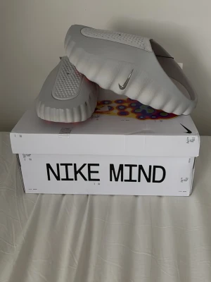 Nike Mind grå tofflor - Nike Mind tofflor i ljusgrått med futuristisk design och vågiga kanter. Tillverkade i ett mjukt, skummaterial med ventilerande hål och en silverfärgad Swoosh på sidan. Sulan har orange detaljer undertill för extra stilpoäng. Passar bättre på för storleken 44