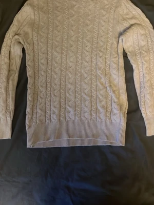 Beige kabelstickad tröja - Kabelstickad tröja i beige med klassiskt mönster och rund hals. Tröjan har långa ärmar och ribbade muddar vid ärmslut och nederkant. Perfekt för lager-på-lager och en stilren look.