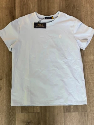 Ljusblå t-shirt från Polo Ralph Lauren - Helt ny ljusblå t-shirt från Polo Ralph Lauren provad en gång men var lite för tajt för mig, jag är 190cm lång och väger 85kg. Storleken  på tröjan är 165-170. Bara att höra av sig vid funderingar! Svarar asap😃