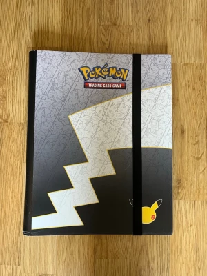 Pokémon samlarkortspärm - Snygg Pokémon samlarkortspärm med Pikachu-motiv och svart elastiskt band. Framsidan har en stor blixt och diskreta Pokémon-figurer i bakgrunden. Perfekt för att organisera och skydda dina Pokémonkort. Officiellt licensierad och tillverkad i tålig plast. Är lite skadad med funkar perfekt för användning. 