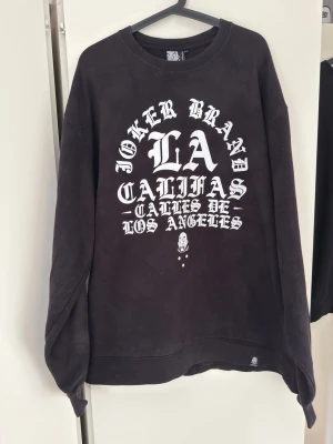 Svart Joker Brand sweatshirt LA-print - Cool svart sweatshirt från Joker Brand med stor vit LA-print och texten 'Joker Brand Calilas Calles De Los Angeles' på bröstet. Tröjan har rund halsringning och avslappnad passform. Perfekt för dig som gillar streetwear och grafiska tryck. Size: xxxl men mera xxl 