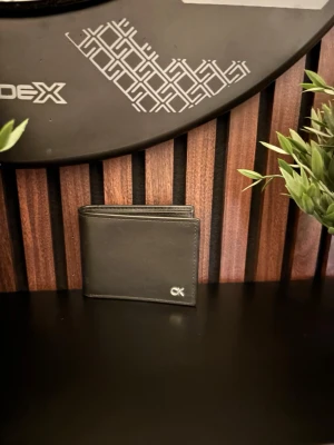 Svart läderplånbok från Calvin Klein - Snygg svart plånbok i äkta läder från Calvin Klein. Klassisk vikbar modell med flera kortfack och diskret CK-logo i metall på framsidan. Perfekt storlek för fickan och stilren design som passar till allt. Helt oanvänd och nypris är 899kr