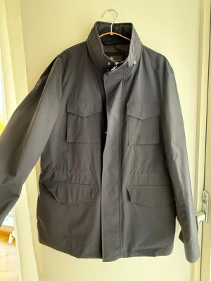 Mörkblå jacka med fyra fickor från Oscar Jacobson - Stilren mörkblå jacka med hög krage och fyra stora fickor framtill. Jackan har dold dragkedja och tryckknappar, samt justerbar midja för bättre passform. Ordinariepris ca 4000kr. Obs  jackan är blå och inte grå, som det kan se ut på bilden. 😊