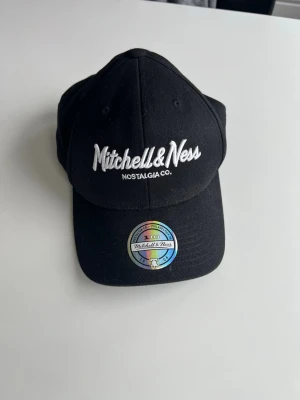 Svart keps från Mitchell & Ness - Snygg svart keps från Mitchell & Ness med broderad logga framtill och bak. Klassisk böjd skärm och justerbar snapback baktill. Tillverkad i bomullsmaterial och har en holografisk etikett på skärmen.