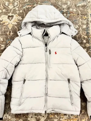 Vit pufferjacka från Ralph Lauren - Snygg vit pufferjacka från Ralph Lauren med huva och dragkedja framtill. Jackan har två sidofickor med dragkedja och den klassiska röda loggan broderad på bröstet. Perfekt för kalla dagar och riktigt stilren look.