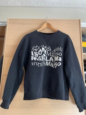 Maison Margiela svart sweatshirt M - Cool svart sweatshirt från Maison Margiela med unik vit textgrafik på bröstet. Tillverkad i Italien av mjuk bomull, perfekt för en avslappnad streetwear-look. Rund halsringning och ribbade muddar vid ärmar och nederkant.