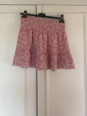 Blommig kortkjol från Gina Tricot - Supersöt blommig kortkjol från Gina Tricot i rosa, vit och grön. Kjolen har smock i midjan för en bekväm passform och ett flowy fall. Perfekt för varma dagar och enkel att matcha med en enkel topp.