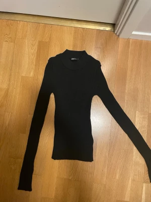 Svart ribbad polotröja Gina Tricot - Tight svart stickad polotröja från Gina Tricot med ribbad struktur. Tröjan har långa ärmar och hög hals, perfekt för lager på lager. Materialet är mjukt och stretchigt, vilket gör den bekväm och enkel att styla till olika outfits. Glittrig 