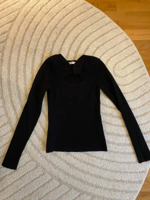 Svart ribbad topp med twistad detalj - Säljer en svart ribbad topp från Samsøe Samsøe med lång ärm och en snygg twistad detalj vid halsen. Toppen har en cut-out framtill som ger en unik look. Materialet är stretchigt och sitter tight mot kroppen. Sitter som på sista bilden! 