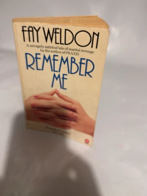 Remember Me av Fay Weldon - Pocketbok med titeln 'Remember Me' av Fay Weldon. Omslaget är beige med blå text och ett foto av två händer. Boken är tryckt på vanligt papper och har en klassisk pocketstorlek, perfekt att ta med sig. En satirisk roman om äktenskap och hämnd.
