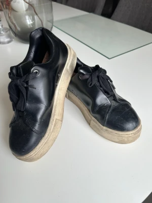 Svarta sneakers från Eytys - Snygga svarta sneakers från Eytys med chunky beige sula och lila hälflik. Skorna har svarta skosnören och är tillverkade i slätt läder. Perfekta för dig som gillar minimalistisk och stilren design.