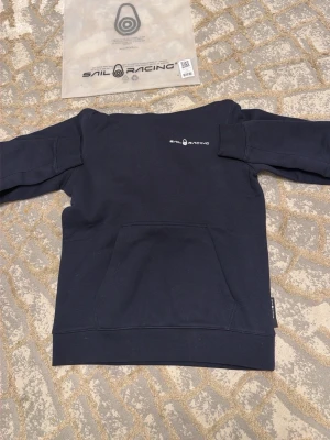 Svart hoodie från Sail Racing - Svart hoodie från Sail Racing med vit logga på bröstet och en stor ficka framtill. Klassisk huva med logga baktill och ribbade muddar. Perfekt för en avslappnad och sportig stil.