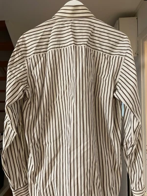 Randig skjorta från Selected - Snygg vit skjorta från Selected med bruna och svarta ränder. Klassisk krage och långärmad modell, perfekt för en clean och stilren look. Tillverkad i mjuk bomull som känns skön mot huden.