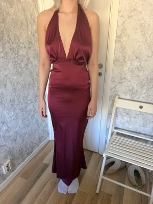 Vinröd långklänning med öppen rygg - Snygg vinröd balklänning i satinliknande material med djup v-ringning och öppen rygg. Klänningen har halterneck och knyt i ryggen för en elegant siluett. Perfekt för dig som vill sticka ut på bal eller fest. Person i fråga är 164-167 cm för referens.