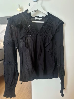 Svart blus med volanger från Neo Noir - Trendig svart blus från Neo Noir med volangdetaljer, v-ringning och broderade hål längs ärmar och bröst. Långärmad modell med smock vid ärmslut och lätt transparent material som ger en luftig känsla. Perfekt för dig som gillar en romantisk och cool stil.